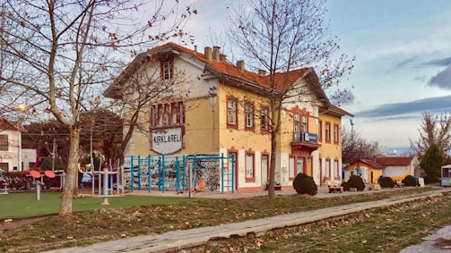A quaint, old-fashioned two-story building with a sign reading 'KIRKLARELI' stands amidst a small, serene park. The building features yellow and red hues, with a tiled roof and multiple windows. Leafless trees surround the area, and there are outdoor exercise equipment and benches nearby. The atmosphere is calm and rustic, with a backdrop of soft clouds in the sky.