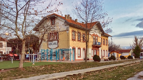 A quaint, old-fashioned two-story building with a sign reading 'KIRKLARELI' stands amidst a small, serene park. The building features yellow and red hues, with a tiled roof and multiple windows. Leafless trees surround the area, and there are outdoor exercise equipment and benches nearby. The atmosphere is calm and rustic, with a backdrop of soft clouds in the sky.