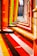 A vibrant urban scene with colorful translucent panels casting vivid red, orange, and yellow shadows on the pavement, creating a mesmerizing pattern. The panels stand alongside a brick building, and the street appears empty with a historic building visible in the background.