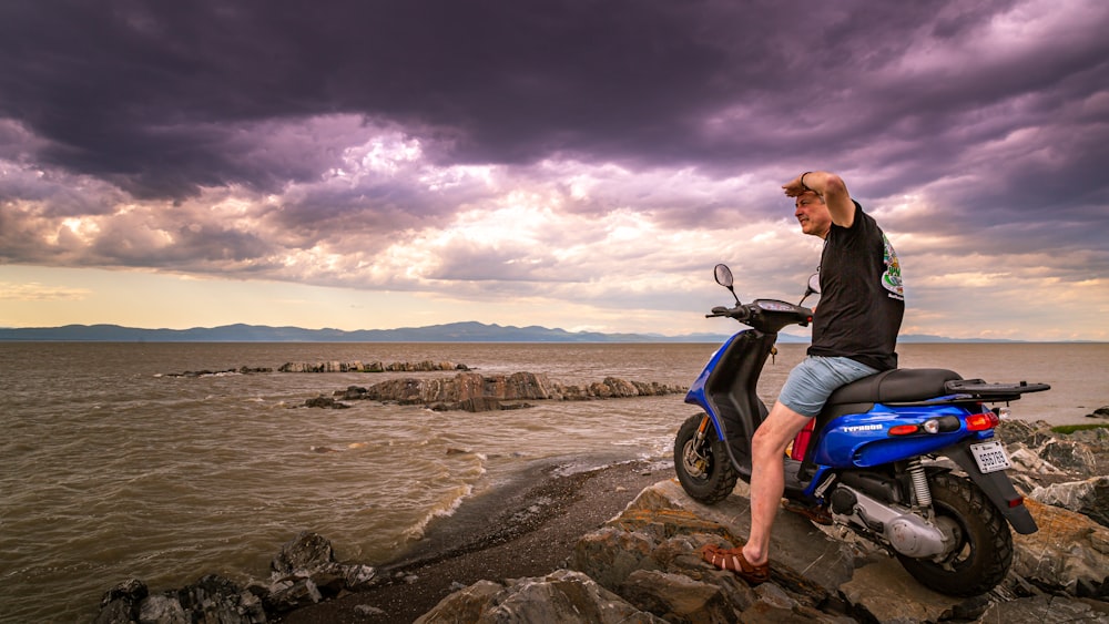 검은 재킷과 파란색 데님 청바지를 입은 남자가 낮 동안 해안에서 오토바이에 앉아 있다