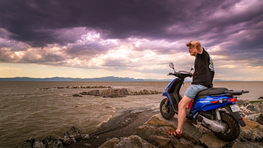 man in black jacket and blue denim jeans sitting on motorcycle on shore during daytime in Rimouski Canada