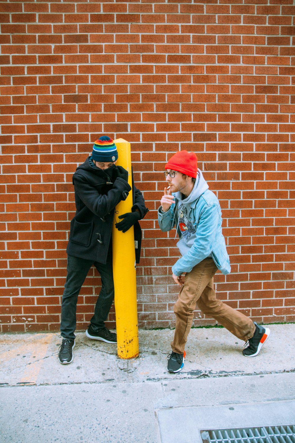 man in blue jacket and brown pants wearing orange knit cap leaning on brown brick wall