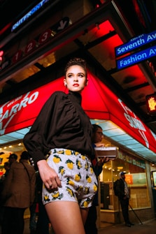 A fashionable person is standing confidently in an urban environment at night, wearing a stylish black blouse and patterned shorts with vibrant fruit designs. The background features a bustling street scene with bright red and blue neon lights, various shop signs, and people walking by, creating a lively city atmosphere.