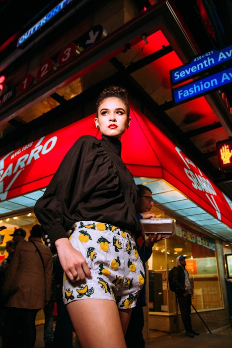 A fashionable person is standing confidently in an urban environment at night, wearing a stylish black blouse and patterned shorts with vibrant fruit designs. The background features a bustling street scene with bright red and blue neon lights, various shop signs, and people walking by, creating a lively city atmosphere.