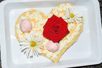 Hands gently holding a heart-shaped cake decorated with soft pink icing and edible flowers
