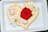 A heart-shaped cake is adorned with creamy frosting and decorated with vibrant flowers. A bright red rose sits prominently in the center, surrounded by two white daisies and two small pink buds.