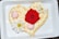 A heart-shaped cake is adorned with creamy frosting and decorated with vibrant flowers. A bright red rose sits prominently in the center, surrounded by two white daisies and two small pink buds.