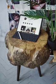 A rustic wooden table with natural textures is topped with a digital tablet that has a keyboard attached, displaying a website. A stylus rests on the table, and the background features magazine clippings and a partial view of a green potted plant.
