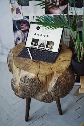 A rustic wooden table with natural textures is topped with a digital tablet that has a keyboard attached, displaying a website. A stylus rests on the table, and the background features magazine clippings and a partial view of a green potted plant.