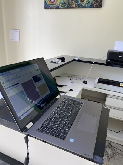 A modern laptop is open on a desk, displaying a web page. The setup includes a printer to the right, other office supplies, and a piece of colorful art on the wall. The desk and walls have a clean and organized appearance.