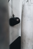 A gym setting featuring a hanging black punching bag attached to a white brick wall and a large white standing punching bag in the foreground.