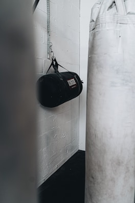 A gym setting featuring a hanging black punching bag attached to a white brick wall and a large white standing punching bag in the foreground.