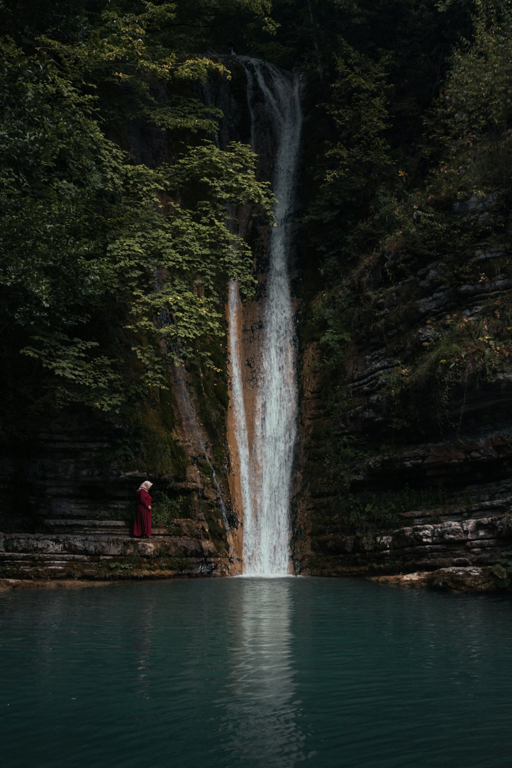 person in red jacket standing on brown wooden dock near waterfalls during daytime