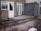 A partially constructed building extension featuring a foundation of bricks and concrete. There are unpainted cinderblock walls and unfinished cement flooring. The existing building has a rough textured exterior with a door and a window.