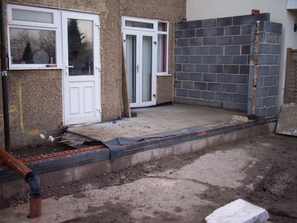 A partially constructed building extension featuring a foundation of bricks and concrete. There are unpainted cinderblock walls and unfinished cement flooring. The existing building has a rough textured exterior with a door and a window.