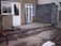 A partially constructed building extension featuring a foundation of bricks and concrete. There are unpainted cinderblock walls and unfinished cement flooring. The existing building has a rough textured exterior with a door and a window.