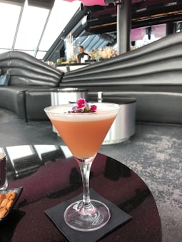 A pink cocktail garnished with small flowers sits on a black napkin on a shiny table. The background features a sleek, modern bar interior with curved black seating and large windows.