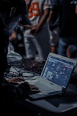 A person is focused on a task involving a laptop with music mixing software. The scene is dimly lit, with a busy background featuring people standing, one wearing a sports jersey with the number 89. Cables are strewn across the table, suggesting an event setup.