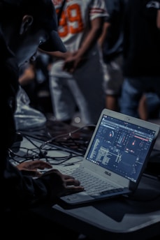 A person is focused on a task involving a laptop with music mixing software. The scene is dimly lit, with a busy background featuring people standing, one wearing a sports jersey with the number 89. Cables are strewn across the table, suggesting an event setup.