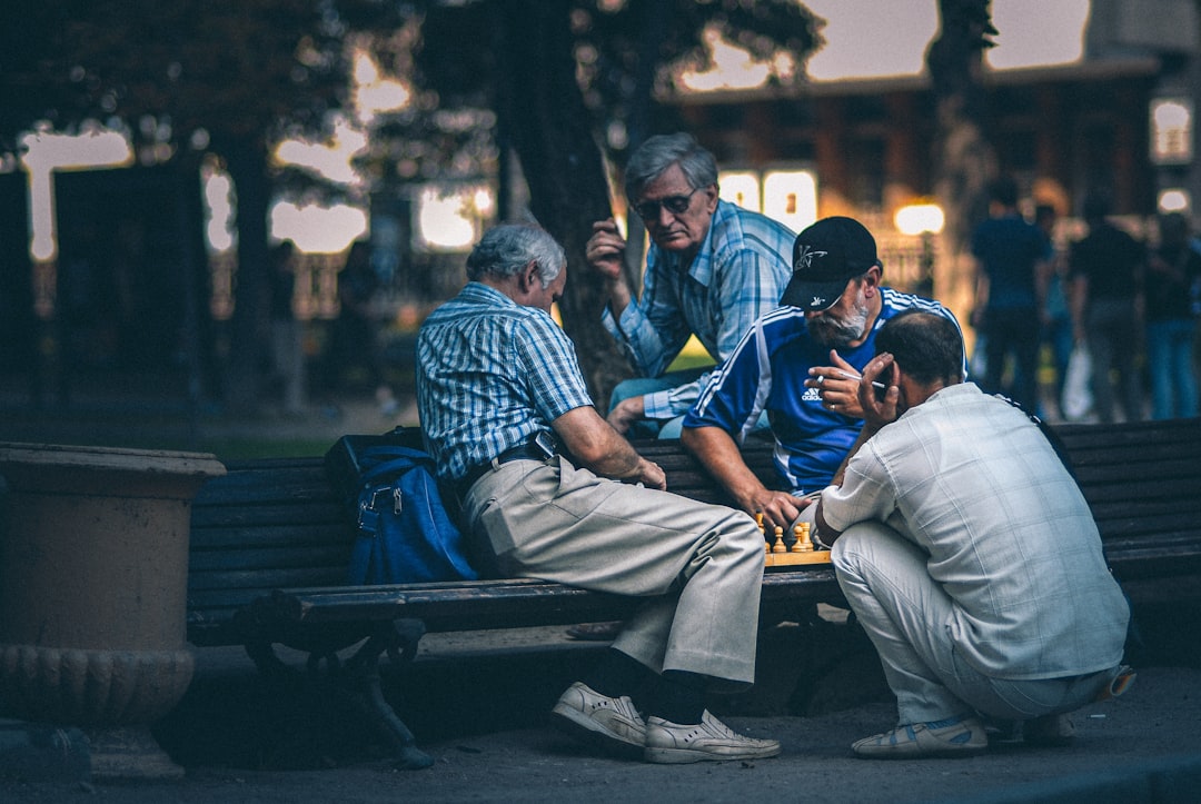man in blue and white plaid dress shirt sitting on bench, Men play chess on the bench in the park.
