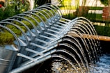 A row of metal watering cans is arranged in a cascading formation, each one tilted forward, allowing water to flow from their spouts in a coordinated, arched pattern into a pond or water feature. The cans are adorned with patches of green moss on their tops, suggesting a blend of nature and man-made design. The setting is a lush garden environment with tall grasses and wooden benches in the background.