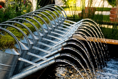 A row of metal watering cans is arranged in a cascading formation, each one tilted forward, allowing water to flow from their spouts in a coordinated, arched pattern into a pond or water feature. The cans are adorned with patches of green moss on their tops, suggesting a blend of nature and man-made design. The setting is a lush garden environment with tall grasses and wooden benches in the background.