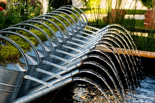 A row of metal watering cans is arranged in a cascading formation, each one tilted forward, allowing water to flow from their spouts in a coordinated, arched pattern into a pond or water feature. The cans are adorned with patches of green moss on their tops, suggesting a blend of nature and man-made design. The setting is a lush garden environment with tall grasses and wooden benches in the background.