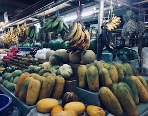 A market stall displaying a variety of fresh fruits, including bunches of bananas hanging from above, pineapples, papayas, melons, and apples arranged neatly on counters. The scene is bustling with diverse and colorful produce in a busy market setting.