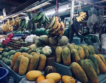 A market stall displaying a variety of fresh fruits, including bunches of bananas hanging from above, pineapples, papayas, melons, and apples arranged neatly on counters. The scene is bustling with diverse and colorful produce in a busy market setting.