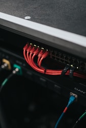 A close-up of a network switch with multiple red Ethernet cables connected. There are also a few black and blue cables plugged into peripheral devices or ports. The area is possibly part of a server room or data center setup.
