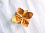 A delicate origami flower crafted from brown paper with five petals, each folded intricately to form a symmetrical pattern. The flower is placed on a plain, textured white surface, creating a simple and elegant contrast.