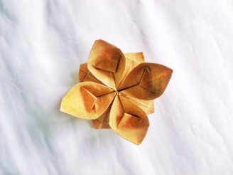 A delicate origami flower crafted from brown paper with five petals, each folded intricately to form a symmetrical pattern. The flower is placed on a plain, textured white surface, creating a simple and elegant contrast.