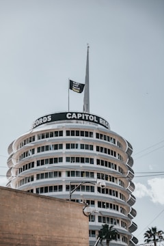 A cylindrical building with multiple floors, featuring the words 'CAPITOL RECORDS' prominently displayed near the top. There is a flagpole with a flag reading 'BLACK LIVES MATTER' extending from the roof. The building has a series of circular balconies and windows. Palm trees and a streetlamp are visible in the foreground.