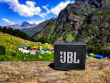 A portable JBL speaker is placed on a rock in a vibrant mountain landscape. The background features lush green hills, colorful tents, and towering mountains under a blue sky with fluffy white clouds.