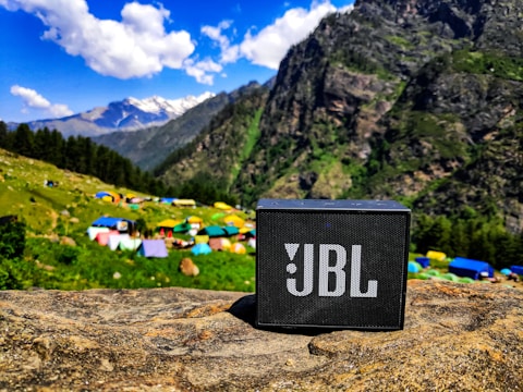 A portable JBL speaker is placed on a rock in a vibrant mountain landscape. The background features lush green hills, colorful tents, and towering mountains under a blue sky with fluffy white clouds.