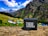 A portable JBL speaker is placed on a rock in a vibrant mountain landscape. The background features lush green hills, colorful tents, and towering mountains under a blue sky with fluffy white clouds.