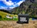 A portable JBL speaker is placed on a rock in a vibrant mountain landscape. The background features lush green hills, colorful tents, and towering mountains under a blue sky with fluffy white clouds.