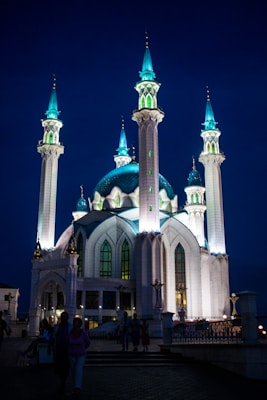 A majestic mosque illuminated against a deep blue evening sky, characterized by its tall, lit minarets and a large central dome with turquoise highlights. The architectural design features intricate details with arched windows and graceful curves. People are visible in shadowed silhouettes near the entrance.