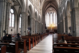 The image depicts the interior of a grand cathedral with high vaulted ceilings and large arched windows. Wooden pews are arranged in rows, leading up to a distant altar adorned with religious statues and paintings. Several people are seated along the pews, suggesting a serene and contemplative atmosphere.