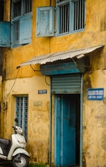 A weathered yellow building with blue shutters and blue entrance door is shown. A white scooter is parked in front, near a sign on the wall reading 'Allied Trades, Jew Town, Kochi-2'. The building has a rustic and slightly rundown appearance, adding to its charm.