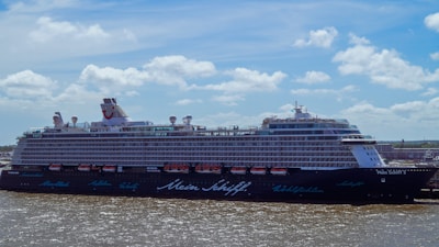 A large cruise ship is docked near a port under a partly cloudy sky. The vessel has multiple decks with rows of windows and lifeboats visible on the sides. The water is slightly choppy, and there are faint outlines of buildings on the horizon.