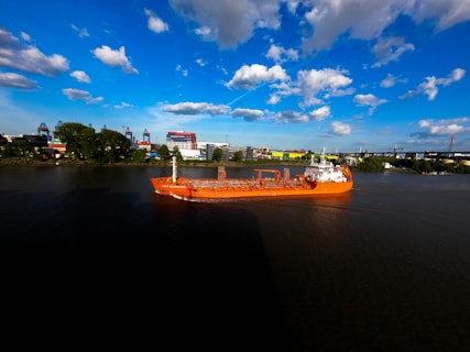 A large orange cargo ship is sailing on a wide river. The background includes a vibrant cityscape with various buildings and cranes visible. The sky is bright with scattered clouds.