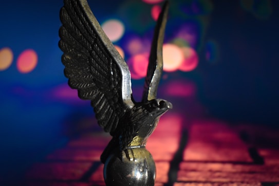 A small, metallic statue of an eagle with wings spread sits prominently, set against a backdrop featuring out-of-focus, colorful lights creating a bokeh effect. The scene gives an impression of depth with vivid hues of blue, pink, and purple.