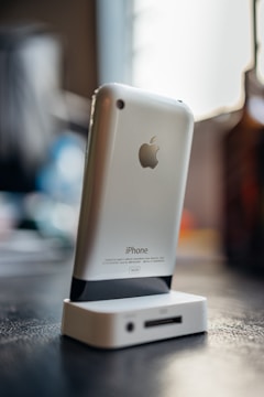 A vintage iPhone with a silver back is docked in an old-style charging station on a dark surface. The background is blurred with soft lighting, indicating a warm ambiance.