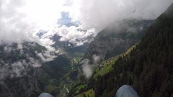 A breathtaking aerial view of a mountainous landscape covered with clouds. Verdant valleys and steep cliffs can be seen, along with a small village nestled among the greenery. Dense forests cover the lower slopes, while the peaks are partially obscured by billowing clouds. The perspective suggests an adventure or paragliding experience.