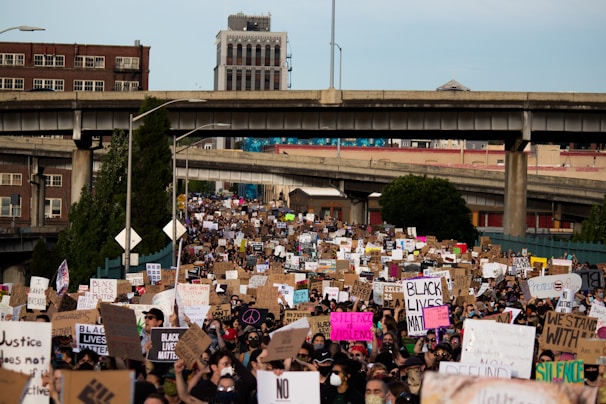 A large crowd of people gathers in a protest, holding signs with slogans related to social justice and equality. The scene takes place on a street with overpasses and buildings in the background. The signs have messages like 'Black Lives Matter' and 'Justice'.