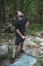 A person is standing in a forested area, wearing outdoor clothing and holding a climbing rope. The individual is looking upward with focused expression, suggesting engagement in an activity like rock climbing or belaying. The surrounding environment is lush with greenery and scattered rocks.
