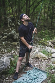 A person is standing in a forested area, wearing outdoor clothing and holding a climbing rope. The individual is looking upward with focused expression, suggesting engagement in an activity like rock climbing or belaying. The surrounding environment is lush with greenery and scattered rocks.