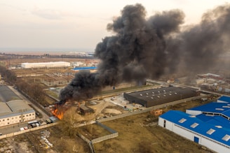 A large plume of thick black smoke rises from a fire at an industrial site. The fire is located near several large buildings with blue and white roofs. Open land and other industrial buildings are visible in the background.