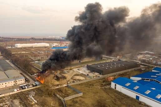 A large plume of thick black smoke rises from a fire at an industrial site. The fire is located near several large buildings with blue and white roofs. Open land and other industrial buildings are visible in the background.